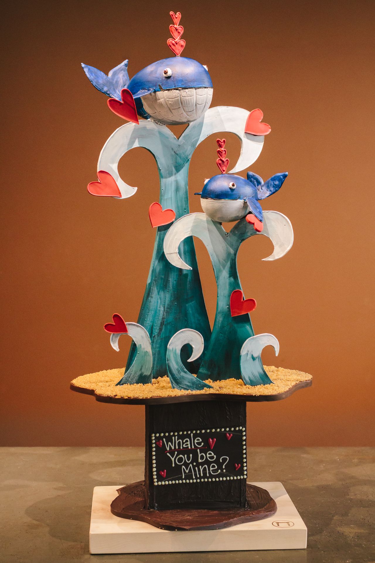 A large chocolate Valentine's sculpture featuring whales on abstract waves. At the bottom is the pun 'Whale You Be Mine?'