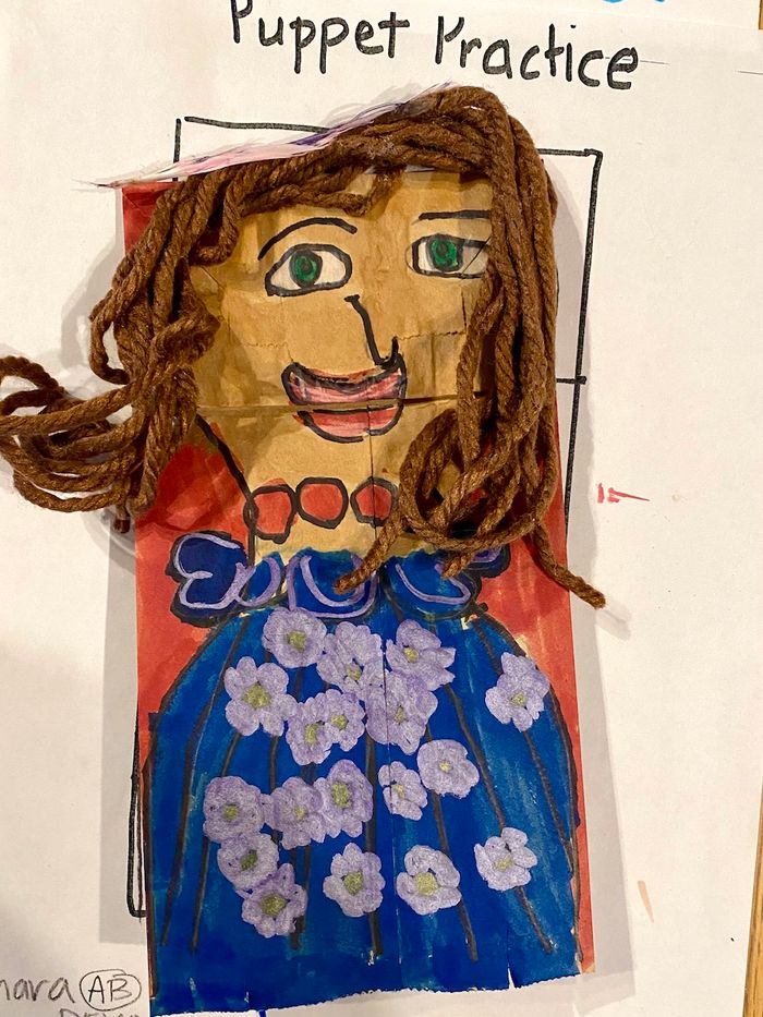 A drawn puppet made out of a brown paper bag. The puppet has brown yarn for hair and is wearing a blue shirt with white flowers on it.
